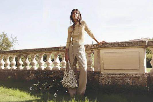 Woman in beige outfit holding a patterned bag, standing against a stone wall with greenery.