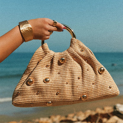 Woven handbag with metallic accents held by a hand against a beach background